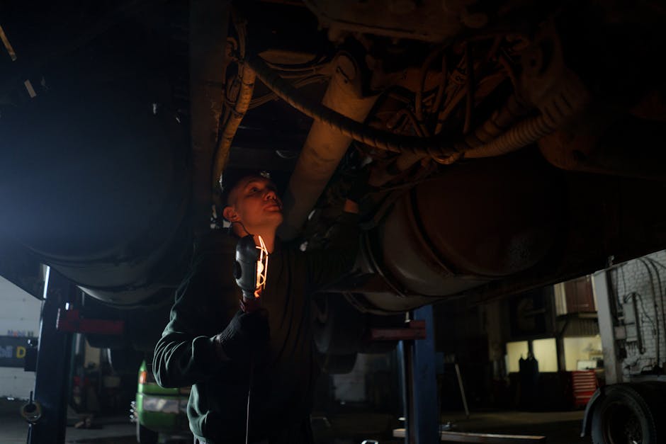 Mechanic examines vehicle underbody in dimly lit workshop, holding inspection light.